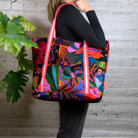 A person holding a large tote bag with vibrant floral artwork and neon pink cord handles.