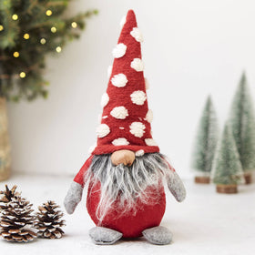 A fabric jolly gonk decoration with a long grey beard and a tall red spotted hat, standing next to a Christmas tree and pine cones.