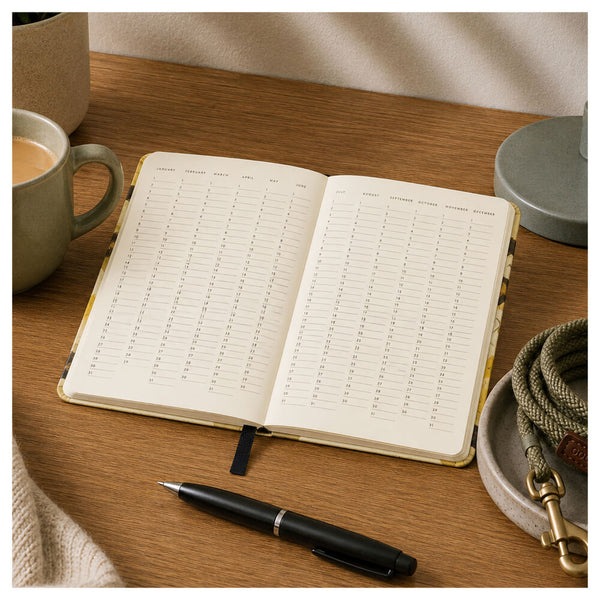 Open book with Months of the year and days of the month on a wooden table next to a mug and pen.