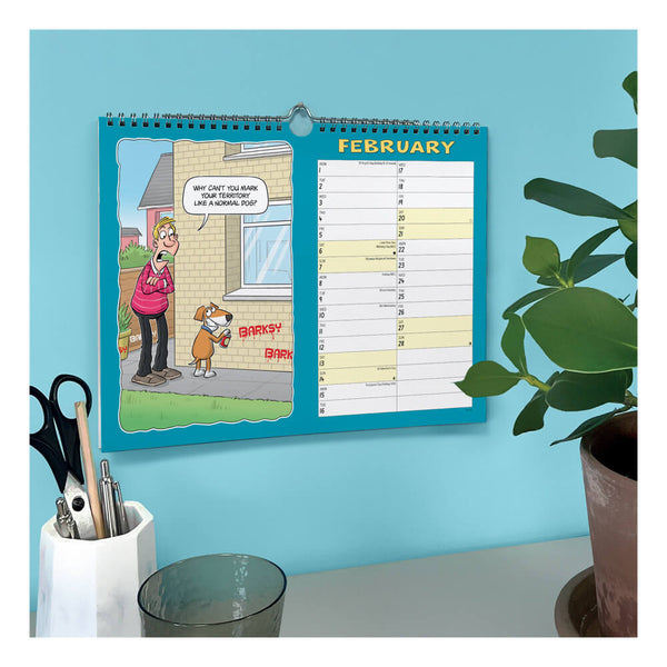 A spiral-bound desk calendar hanging on a light blue wall, showing a cartoon scene on the left and a numbered daily planner on the right. The calendar is styled in a home office setting with a plant, a pen cup, and a glass on a white surface below.