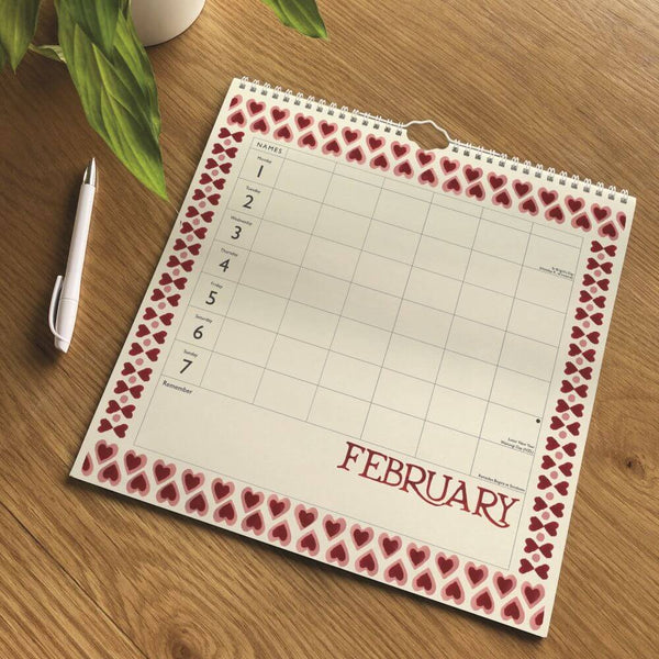 A spiral-bound February calendar page with a red and pink heart pattern border, shown on a wooden tabletop next to a white pen and a green plant.
