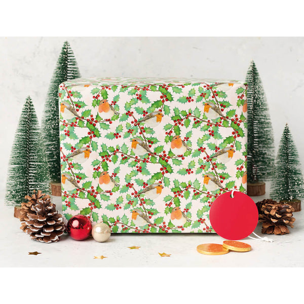 A square gift box wrapped in festive Christmas paper with a repeating pattern of green holly leaves, red berries, and orange birds on an off-white background. The box is styled with miniature green Christmas trees, pinecones, red and gold ornaments, and a red ornament tag in front of it, set against a light gray marble background.