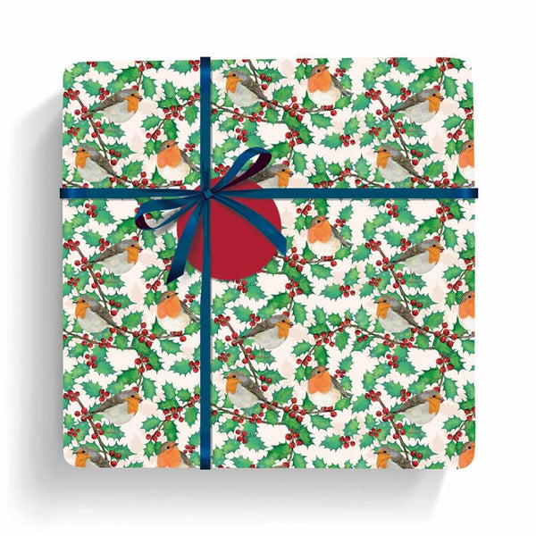 A square gift box wrapped in a festive Christmas pattern featuring green holly leaves, red berries, and orange birds on a white background, tied with a dark blue ribbon and bow with a red spherical ornament in the center, shown on a white background.