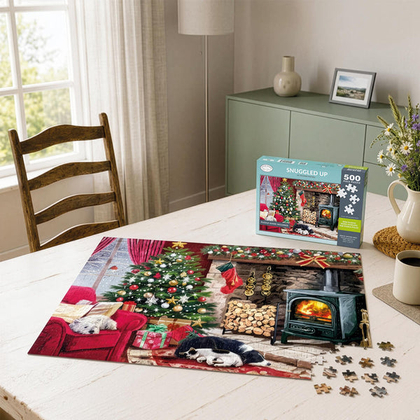 A completed Christmas-themed jigsaw puzzle and its box sit on a white table in a cozy room setting, with loose puzzle pieces scattered nearby.