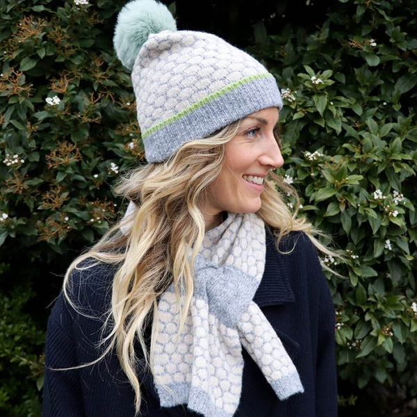 A woman wearing a gray and mint green popcorn knit hat with a pom-pom and a matching scarf. She is standing in front of a bush.