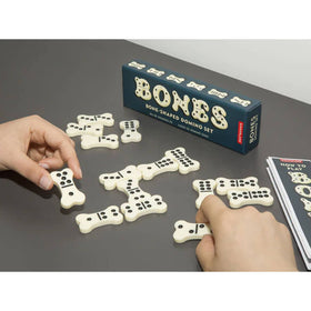 A person's hands setting up a game of Bones Dominoes on a table, with pieces shaped like bones and the game's box in the background.