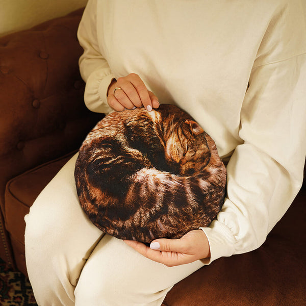 A person in cream sweatpants holds a round plush pillow shaped like a curled-up brown tabby cat hot water bottle, resting on a brown upholstered chair.