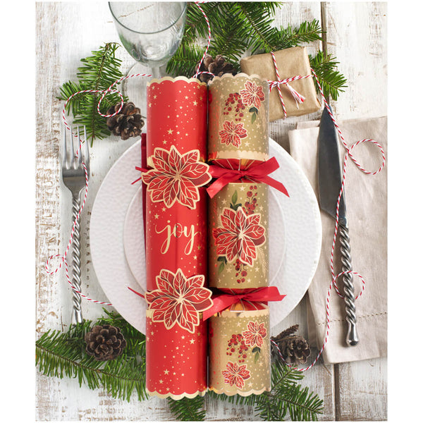 Two Christmas-themed cylindrical crackers wrapped in red and gold paper with floral designs and red ribbons, arranged on a white plate with a glass, fork, knife, pinecones, and greenery on a whitewashed wooden table.