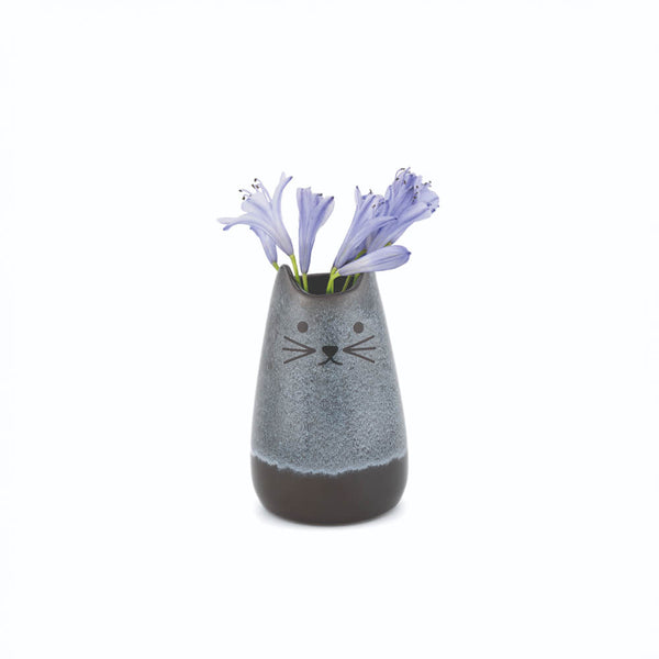 A small ceramic cat-shaped vase with a gray and dark brown glaze, featuring a simple black cat face design, holding three light purple flowers against a plain white background.