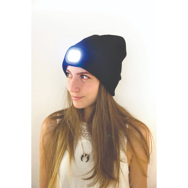A woman wearing a black beanie with an integrated LED headlamp that is turned on, emitting a bright white light. The background is a plain off-white wall.
