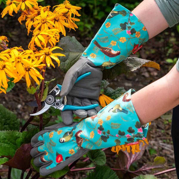 Person wearing floral gardening gloves holding garden shears in a garden setting with yellow flowers.