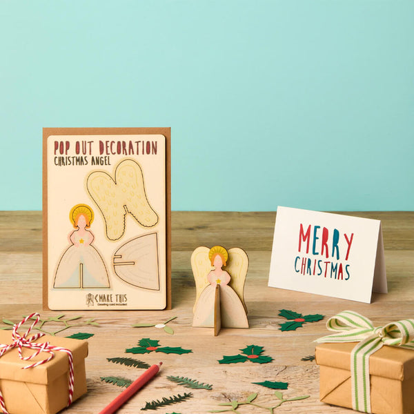 Pop-out Christmas angel decoration with packaging and 'Merry Christmas' card on a wooden surface.