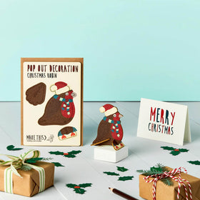 A pop-out Christmas card featuring a festive robin decoration in a Santa hat and striped scarf, displayed on a stand with a 'Merry Christmas' greeting card and a brown envelope, set against a light blue background.