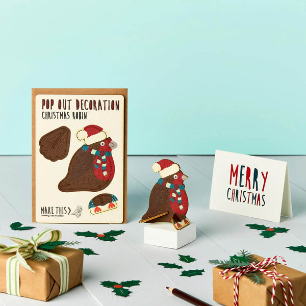 A pop-out Christmas card featuring a festive robin decoration in a Santa hat and striped scarf, displayed on a stand with a 'Merry Christmas' greeting card and a brown envelope, set against a light blue background.
