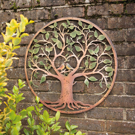 A rustic metal wall plaque depicting a tree with heart-shaped branches and two birds nestled among the leaves, displayed against a brick wall backdrop.