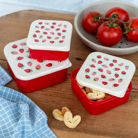 Red and white food containers with strawberry pattern lids on a wooden surface with tomatoes and cookies.