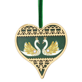 A heart-shaped wooden Christmas ornament featuring two swans facing each other on a dark green background, with a gold-colored laser-cut lace border and a green hanging ribbon.