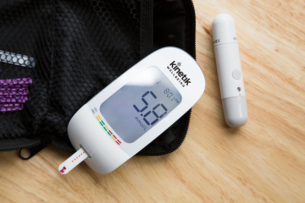 The Kinetik Wellbeing blood glucose meter placed on a table alongside the lancing device and travel pouch that comes with the Blood Glucose Monitoring System.