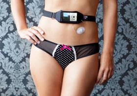 Person wearing black HID-In Multiway Body Band with clear window, shown securing a visible medical device for diabetes management on the abdomen